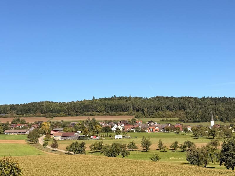 Ein kleines Dorf mit verstreuten Häusern und einer Kirche liegt eingebettet in eine grüne, landwirtschaftlich genutzte Landschaft, umgeben von Feldern und Wäldern unter einem klaren blauen Himmel.