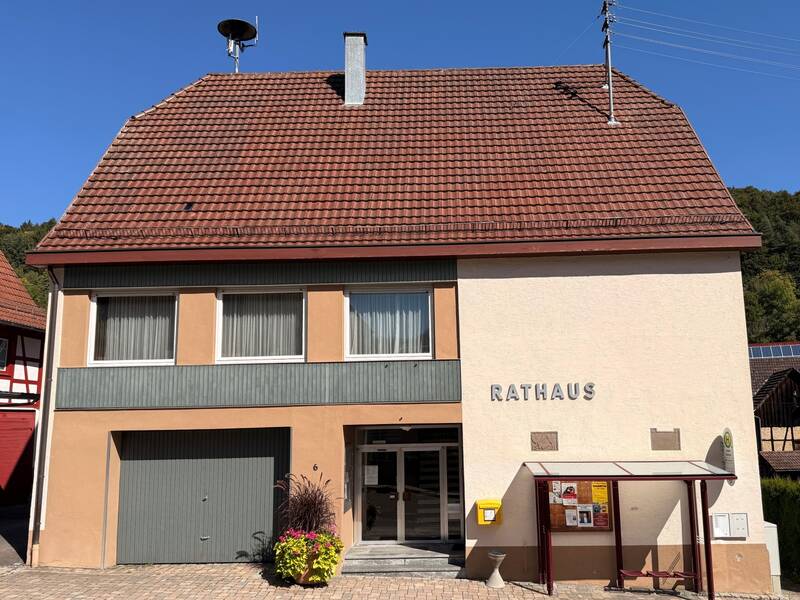 Das Bild zeigt ein Gebäude mit der Aufschrift "Rathaus", einem Eingangsbereich und einem Wartehäuschen rechts im Vordergrund.