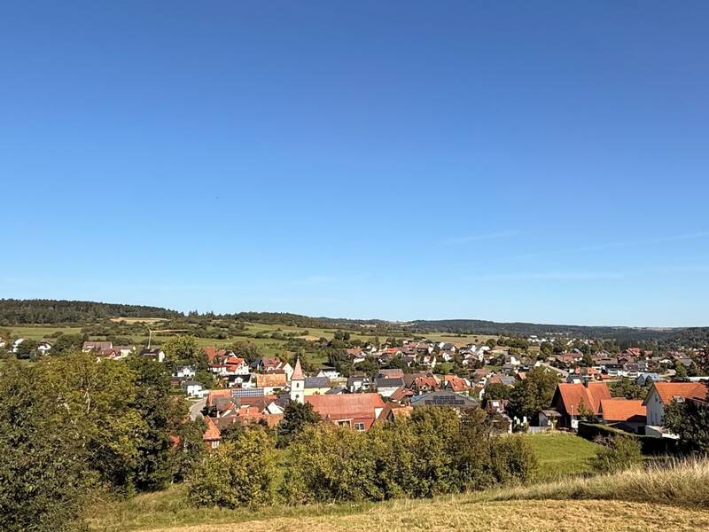 Ein Dorf mit roten Dächern liegt eingebettet in eine grüne Landschaft unter einem klaren blauen Himmel.