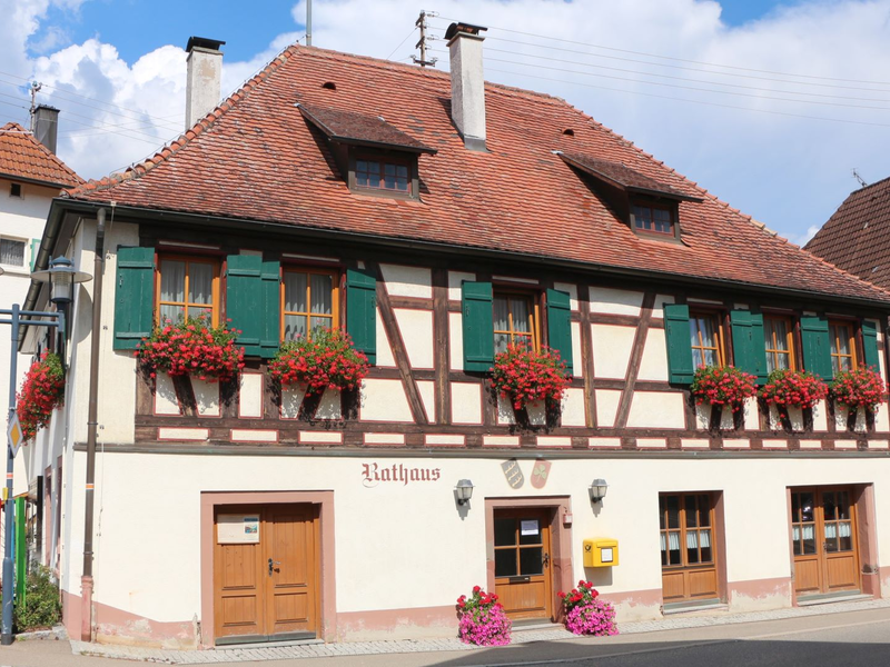 Das Bild zeigt ein Fachwerkhaus mit roten Dachziegeln, grünen Fensterläden und blühenden Blumenkästen, auf dem "Rathaus" steht.