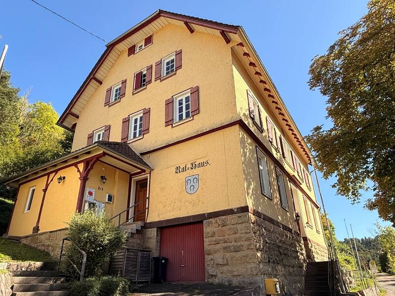 Das Bild zeigt ein großes, gelbes Haus mit roten Fensterläden und einem steinernen Sockel in einer ländlichen Umgebung unter einem klaren, blauen Himmel.