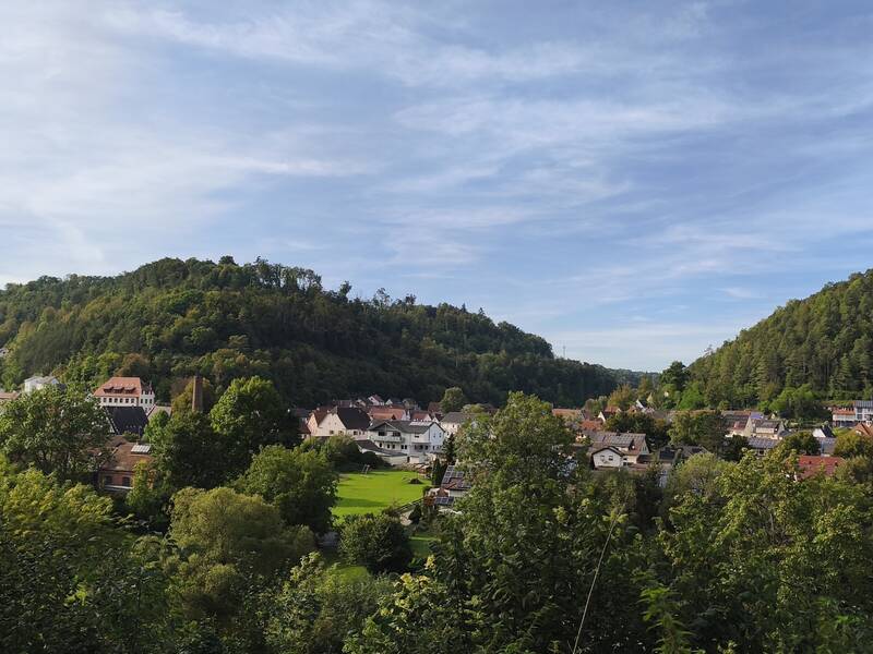Eine idyllische Kleinstadt liegt eingebettet zwischen grünen, bewaldeten Hügeln unter einem leicht bewölkten Himmel.