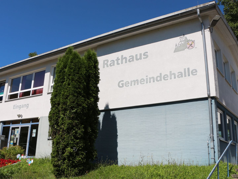 Das Bild zeigt ein zweistöckiges Gebäude mit der Aufschrift "Rathaus Gemeindehalle" und einem Eingangsbereich, umgeben von Grünflächen.