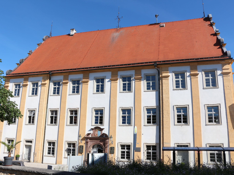 Das Bild zeigt ein historisches Gebäude mit einem roten Ziegeldach und mehreren symmetrisch angeordneten Fenstern an der Fassade.