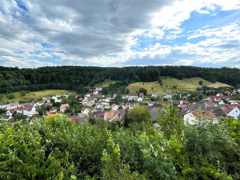 Eine Kleinstadt mit roten und weißen Häusern liegt inmitten einer von Bäumen und grünen Hügeln umgebenen Landschaft unter einem blauen Himmel mit Wolken.