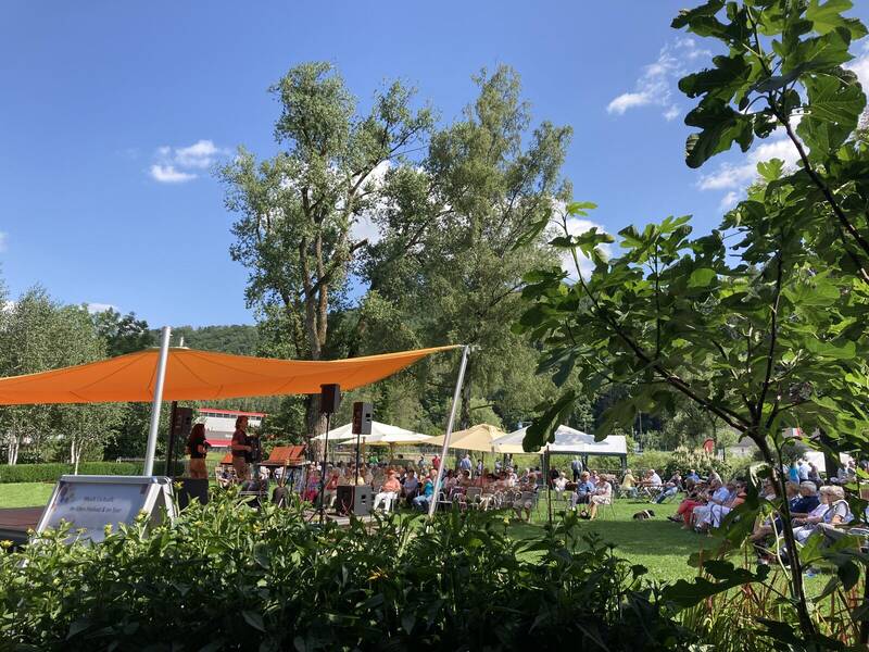 Eine Outdoor-Veranstaltung mit einer Bühne links, auf der eine Band spielt, und vielen Menschen, die unter Sonnenschirmen auf der Wiese sitzen.