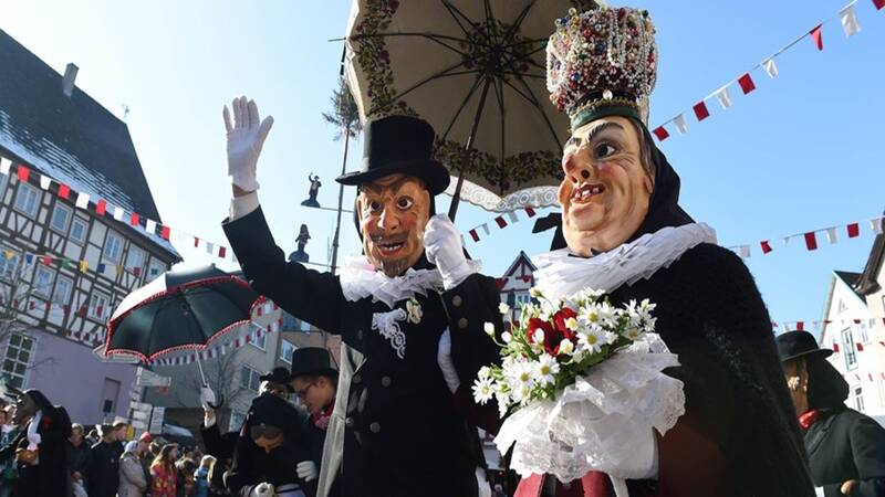Auf dem Bild sind zwei Personen in traditionellen Karnevalskostümen und großen Masken zu sehen, die bei einem Umzug eine Parade genießen, wobei eine Person einen Blumenstrauß hält.