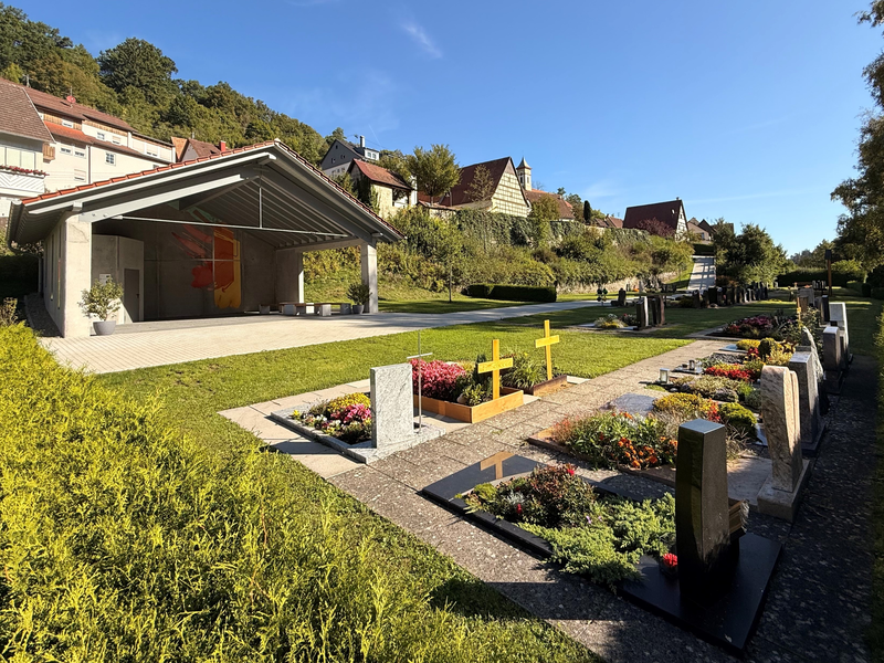 Ein Friedhof mit bunten Blumen, Grabsteinen und einer ruhigen Wegesbiegung inmitten grüner Bäume.