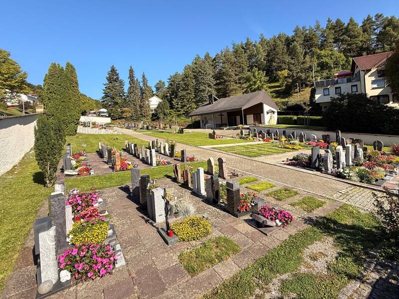 Ein Friedhof mit gepflegten Gräbern und blühenden Blumen ist von Bäumen und einigen Gebäuden umgeben.