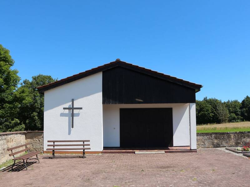 Das Bild zeigt eine kleine, schlichte Friedhofskapelle mit einem Kreuz an der Fassade, umgeben von Bäumen und einem blauen Himmel.