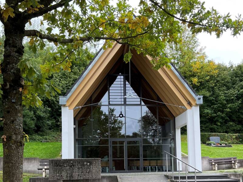 Eine Aussegnungshalle mit einem großen Glasgiebel und einem Spitzdach steht von Bäumen und Grünflächen umgeben.