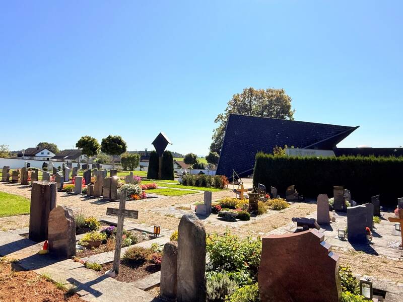 Ein sonniger Friedhof mit Grabsteinen in verschiedenen Formen und Größen, umgeben von grünen Bäumen und einer modernen, geometrisch gestalteten Kapelle im Hintergrund.