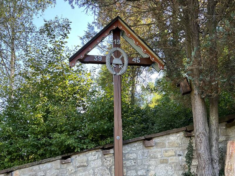 Das Bild zeigt ein hölzernes Friedhofskreuz mit der Aufschrift "1939-1945" vor einer bewaldeten Kulisse und einer Steinmauer.