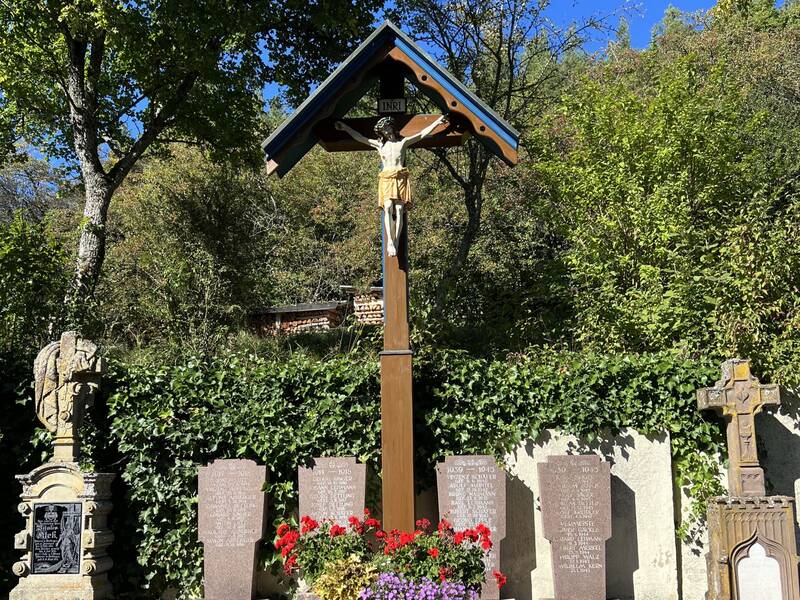 Das Bild zeigt ein hölzernes Friedhofskreuz  vor einer bewaldeten Kulisse.