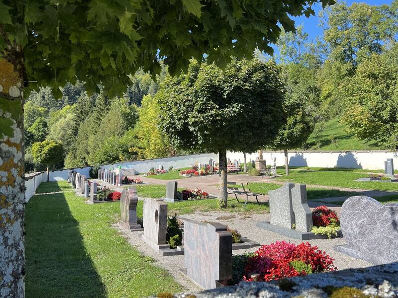 Ein Friedhof mit Grabsteinen und Blumen, umgeben von Bäumen unter einem teilweise bewölkten blauen Himmel.