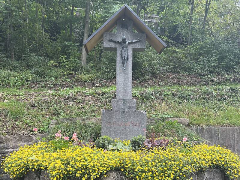 Ein Friedhofskreuz aus Stein mit gelben Blumen vor einem bewaldeten Hintergrund.
