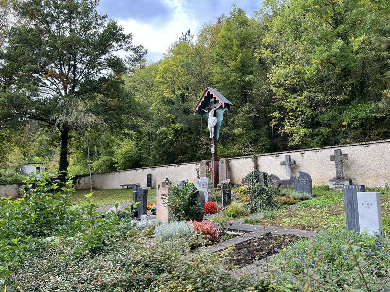 Ein idyllischer Friedhof mit einem großen Kruzifix in der Mitte, umgeben von Grabsteinen und üppiger Vegetation vor einer mit Bäumen bewachsenen Mauer.