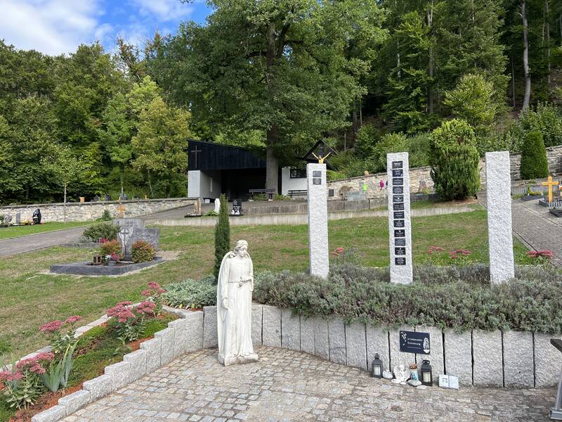 Ein gepflegter Friedhof mit einer Christusstatue, Blumenbeeten, drei Steinsäulen mit Namenstafeln und einem kleinen Gebäude im Hintergrund ist von grünem Wald umgeben.