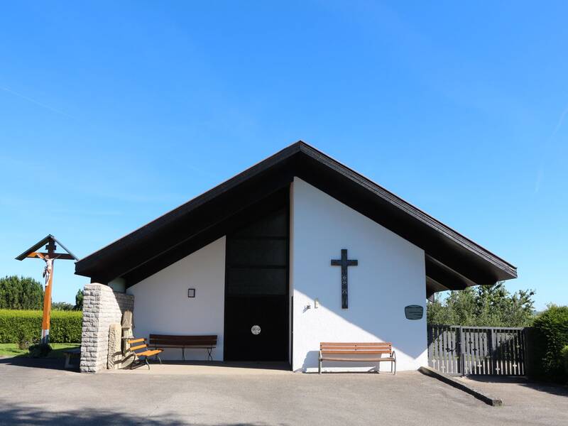 Ein kleines, weißes Gebäude mit schrägem Dach und einem Kreuz an der Vorderseite steht neben einer überdachten Sitzbank und einem Kreuz im Freien.