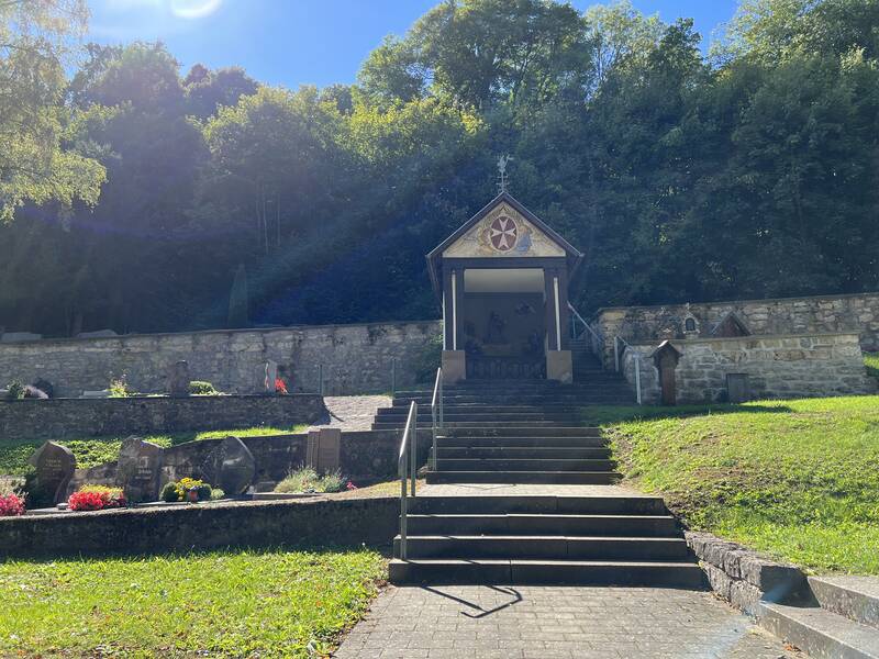 Das Bild zeigt eine kleine Kapelle auf einem Friedhof, umgeben von grüner Vegetation und einer Steinmauer, mit einem Weg und Stufen, die hinaufführen.