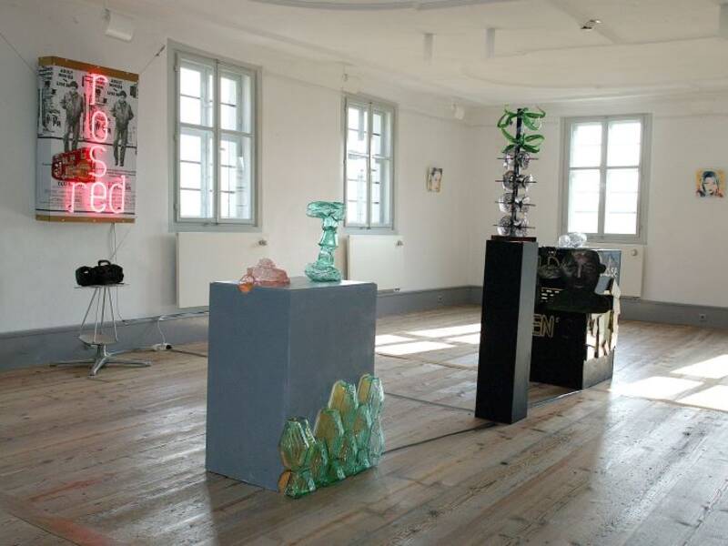 Das Bild zeigt eine Kunstausstellung in einem Raum mit Fenstern und Parkettboden.