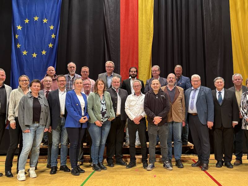 Eine Gruppe von etwa 25 Personen steht in einem Raum vor großen EU- und deutschen Flaggen und posiert für ein Gruppenfoto.