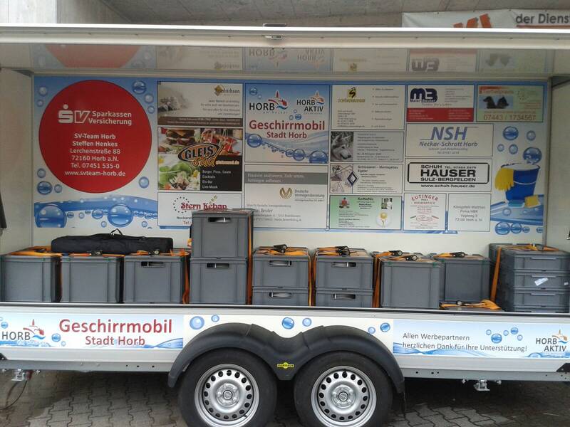 Ein Anhänger mit der Beschriftung "Geschirrmobil Stadt Horb" zeigt mehrere Werbetafeln und gestapelte graue Kisten.