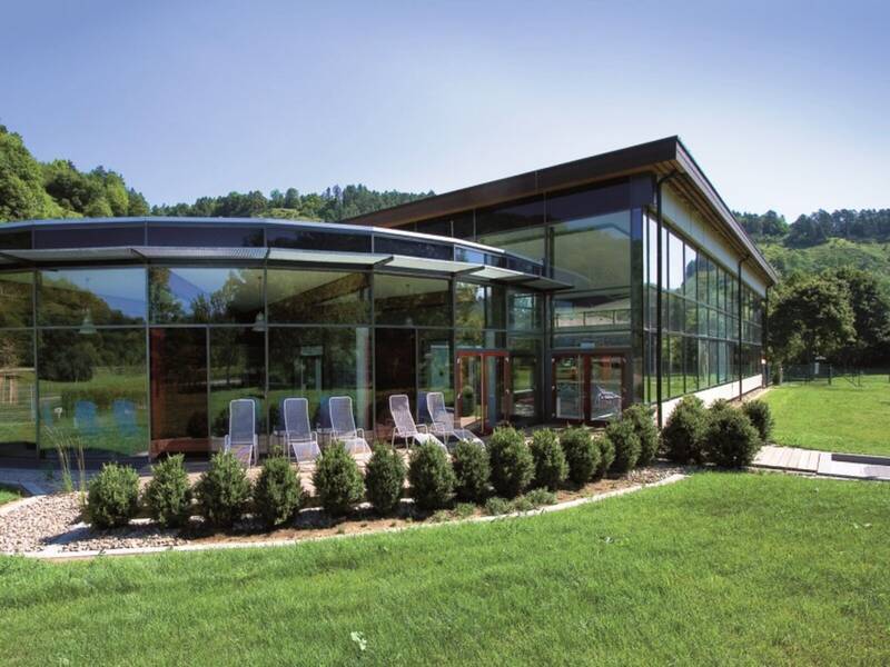 Ein Gebäude mit einer Glasfront. Davor stehen ein paar Liegestühle. Das Gebäude ist umgeben von einer grünen Wiese, Bäumen und Hecken.