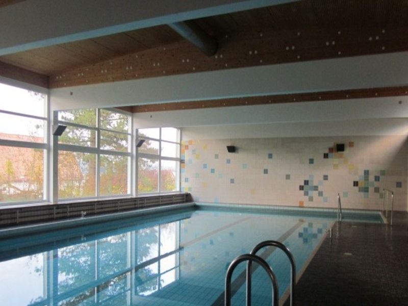 Auf dem Bild ist ein Hallen-Schwimmbad zu sehen mit gefliester Wand und Boden und Fenstern.
