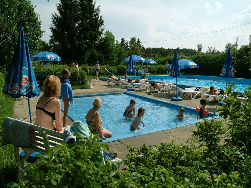 Auf dem Bild ist ein Freibad mit zwei Schwimmbecken zu sehen, in denen mehrere Personen baden. Außerdem gibt es Sonnenschirme und Liegestühle. Das Freibad ist umgeben von grünen Bäumen und Hecken, Es sind
