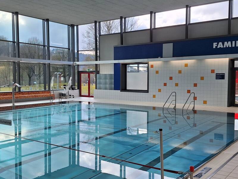 Das Bild zeigt ein Hallenbad mit einem klaren, rechteckigen Pool, umgeben von großen Fenstern und Fliesen, mit einem Schild "Familienbereich" an der Wand.