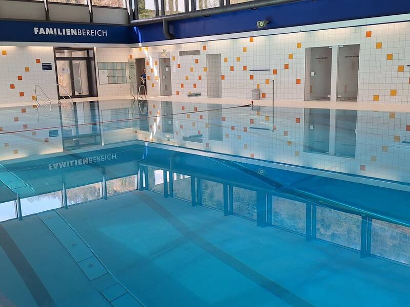 Das Bild zeigt ein Hallenbad mit einem großen, leeren Schwimmbecken und einer gekachelten Wand mit bunten Akzenten, auf der "Famillienbereich" steht.