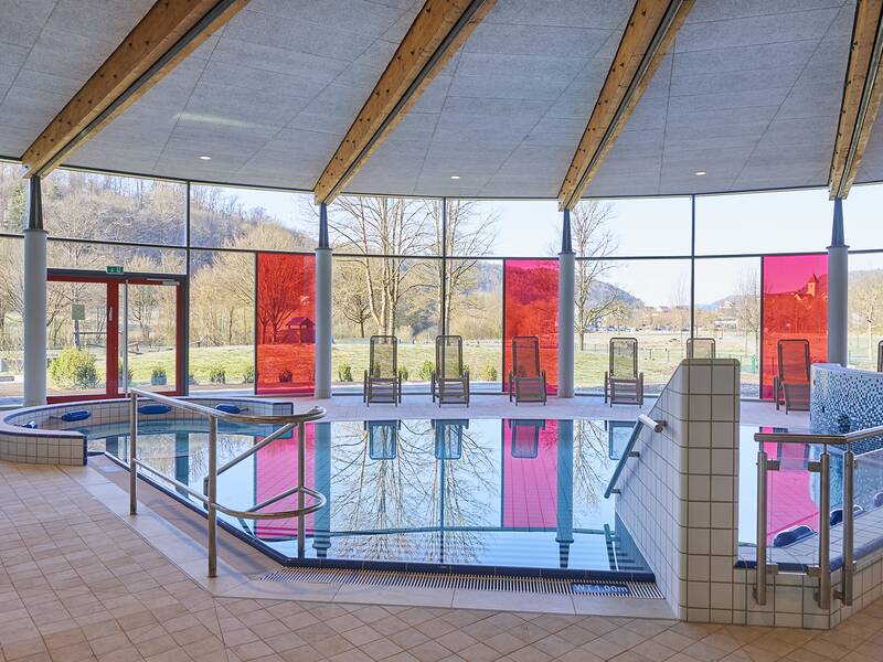 Ein Innenpoolbereich mit Stufen und Handlauf führt in ein rundes Becken, umgeben von großen Fenstern mit roten Elementen, die einen Blick nach draußen bieten.