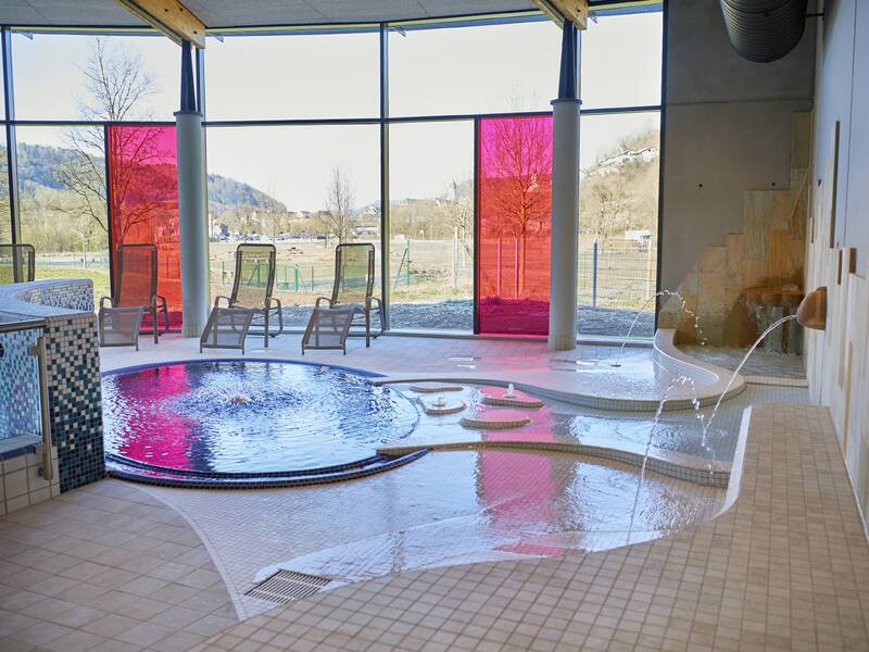 Ein Wellnessbereich mit einem runden Pool und sprudelndem Wasser, umgeben von Liegestühlen vor großen Fenstern mit Ausblick ins Freie.