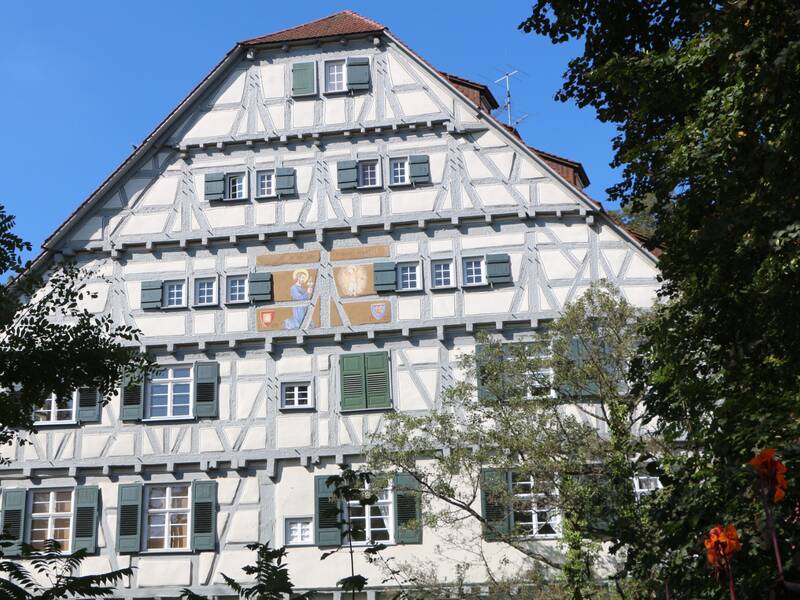Ein helles Fachwerkhaus mit grünen Fensterläden umgeben von Bäumen und Sträuchern.