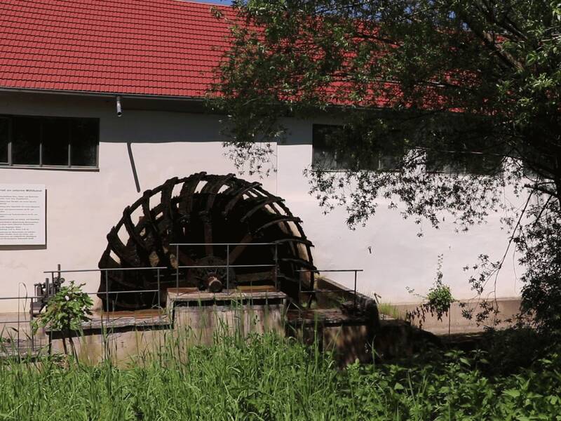 Ein hölzernes Wasserrad und ein Gebäude mit roten Ziegeln umgeben von einer grünen Wiese und Bäumen.