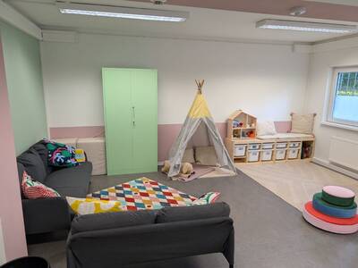 Ein gemütlich eingerichteter Raum mit grauem Sofa, bunten Kissen, einem farbenfrohen Teppich, einem hellen Spielzelt, Regalen mit Kisten und bunten Sitzkissen in einer Kinderspielecke.