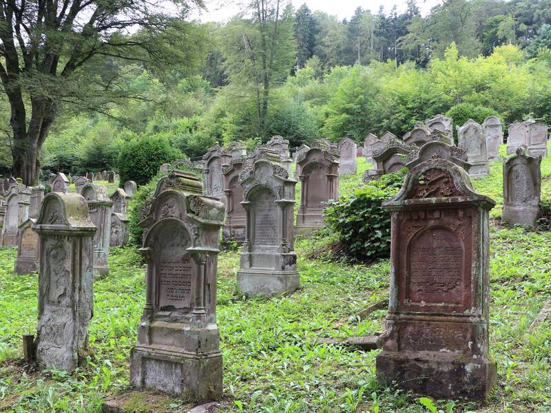 Eine alte, überwachsene Friedhofslandschaft mit vielen verwitterten Grabsteinen, umgeben von dichtem Wald.