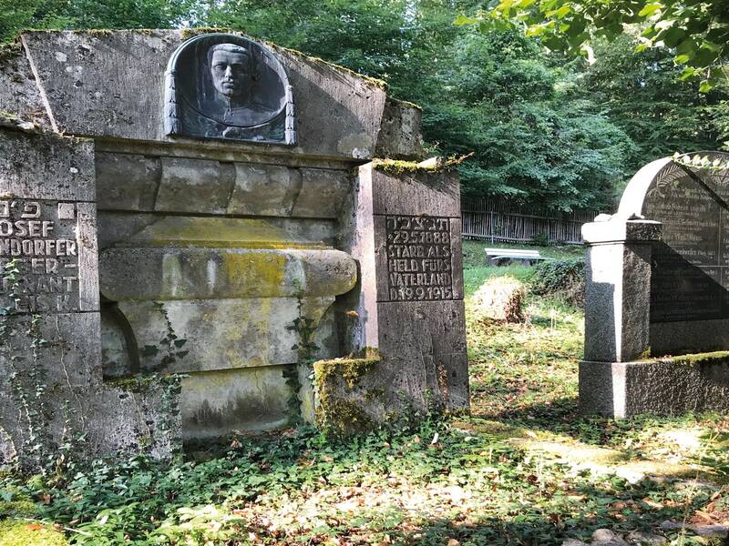 Zwei verwitterte Grabsteine, teilweise mit Moos bedeckt, stehen auf einem bewachsenen Friedhofsgelände im Wald.