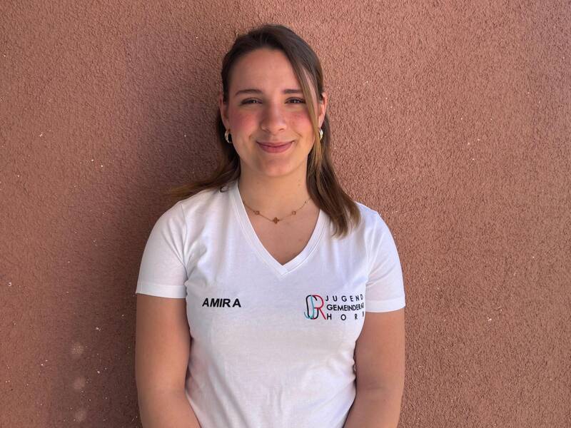 Eine lächelnde Person mit einem weißen T-Shirt, auf dem "AMIRA" und "JUGEND GEMEINDERAT HORN" steht, steht vor einer braunen Wand.