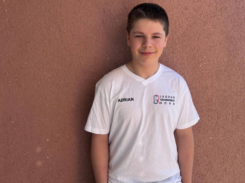 Eine Person in einem weißen T-Shirt mit einem Aufdruck steht vor einer rosafarbenen Wand und lächelt in die Kamera.