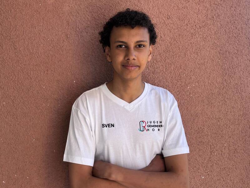 Eine Person mit lockigem Haar steht vor einer braunen Wand und trägt ein weißes T-Shirt mit der Aufschrift "SVEN" und einem Logo.