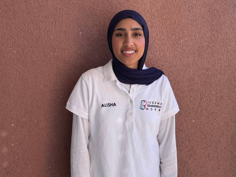 Eine Person mit dunkelblauem Kopftuch und weißem Polohemd, auf dem "ALISHA" und "Jugendgemeinderat Horb" steht, lächelt vor einem rötlichen Hintergrund.