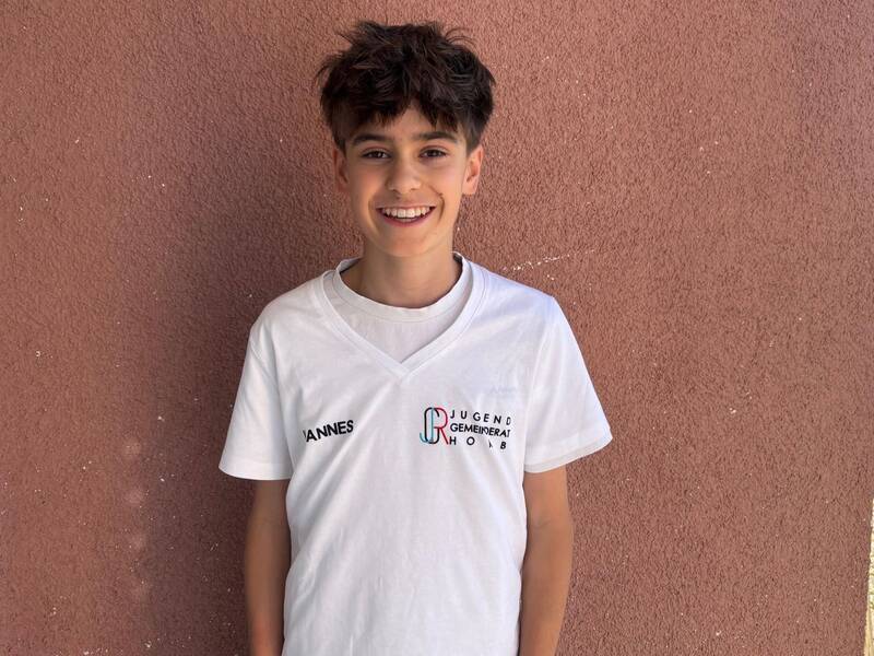 Ein Junge mit braunen Haaren lächelt in einem weißen T-Shirt vor einer rauen, rot-braunen Wand.