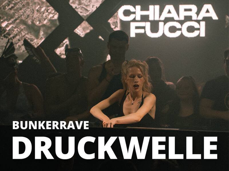Eine Person steht hinter einem DJ-Pult in einem dunklen Raum mit der Aufschrift "Chiara Fucci" und "Bunkerrave Druckwelle" auf der oberen und unteren Bildhälfte.
