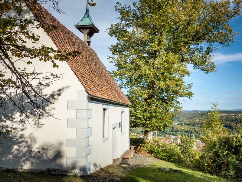 Die Aufnahme zeigt eine kleine weiße Kapelle mit einem roten Ziegeldach und Turm neben einem großen Baum, umgeben von einer idyllischen Landschaft im Hintergrund.