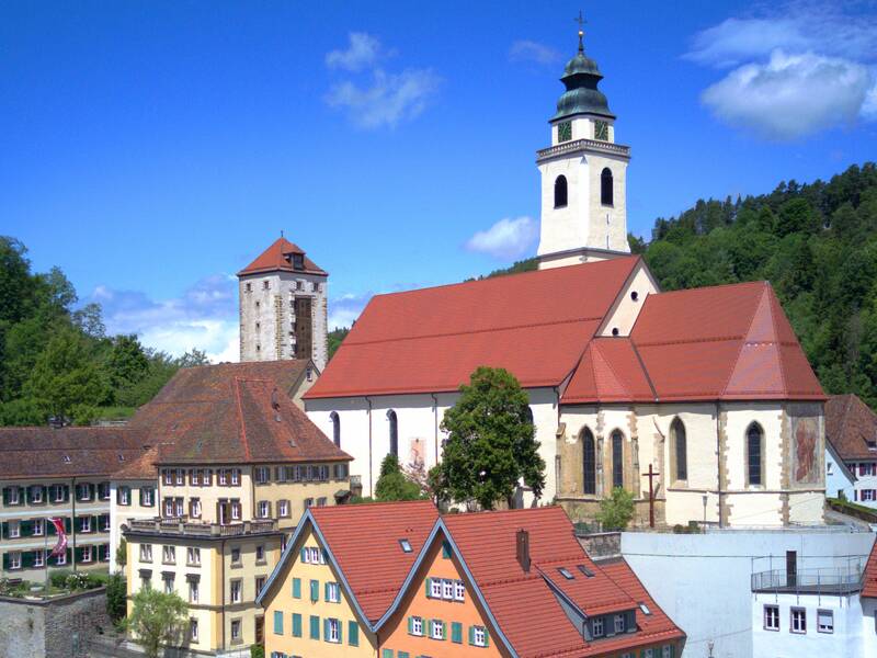 Das Bild zeigt eine Kirche mit rotem Ziegeldach umgeben von mehreren bunten Gebäuden. Im Hintergrund sind Bäume und ein klarer blauer Himmel zu sehen.