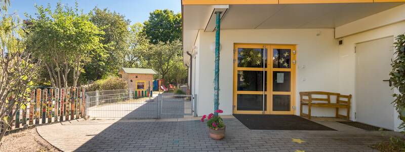 Rückseite des Gebäudes mit Zugang zum Außenspielbereich gepflastert und umzäunt. Im Hintergrund steht ein Spielhaus und ist von Bäumen umgeben.