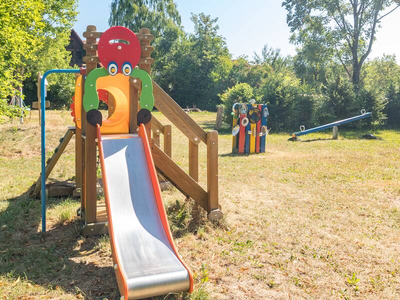 Kleiner Spielturm mit Rutsche und Wippe im Hintergrund von Bäumen eingegezäunt.