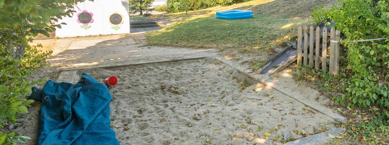 Sandkiste im Vordergrund mit Planschbecken auf Wiese und dem Kindergartengebäude auf der rechten Seite im Bild.